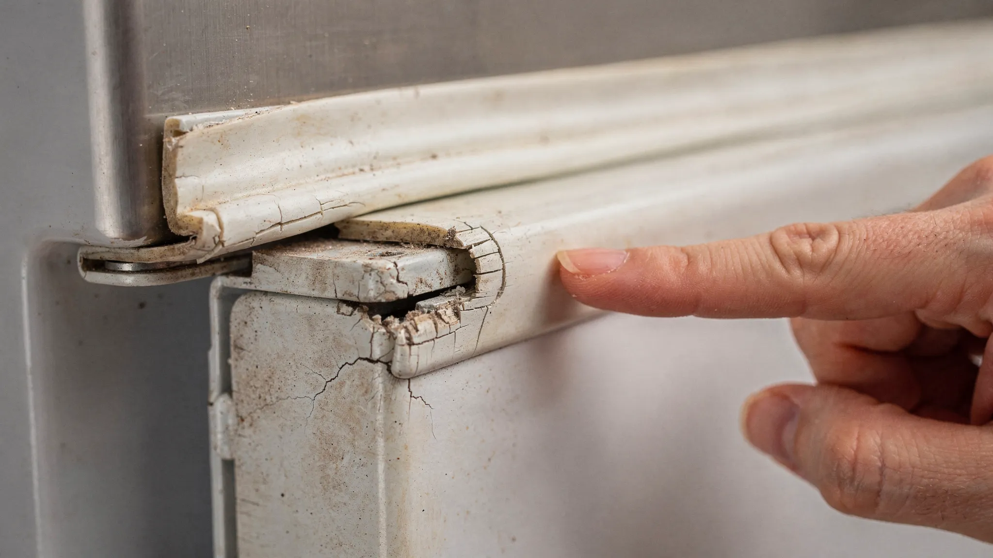 Close-up of a refrigerator door gasket with visible cracks at the corner and slight gaps where the seal meets the frame; a hand points to the damaged area for emphasis.