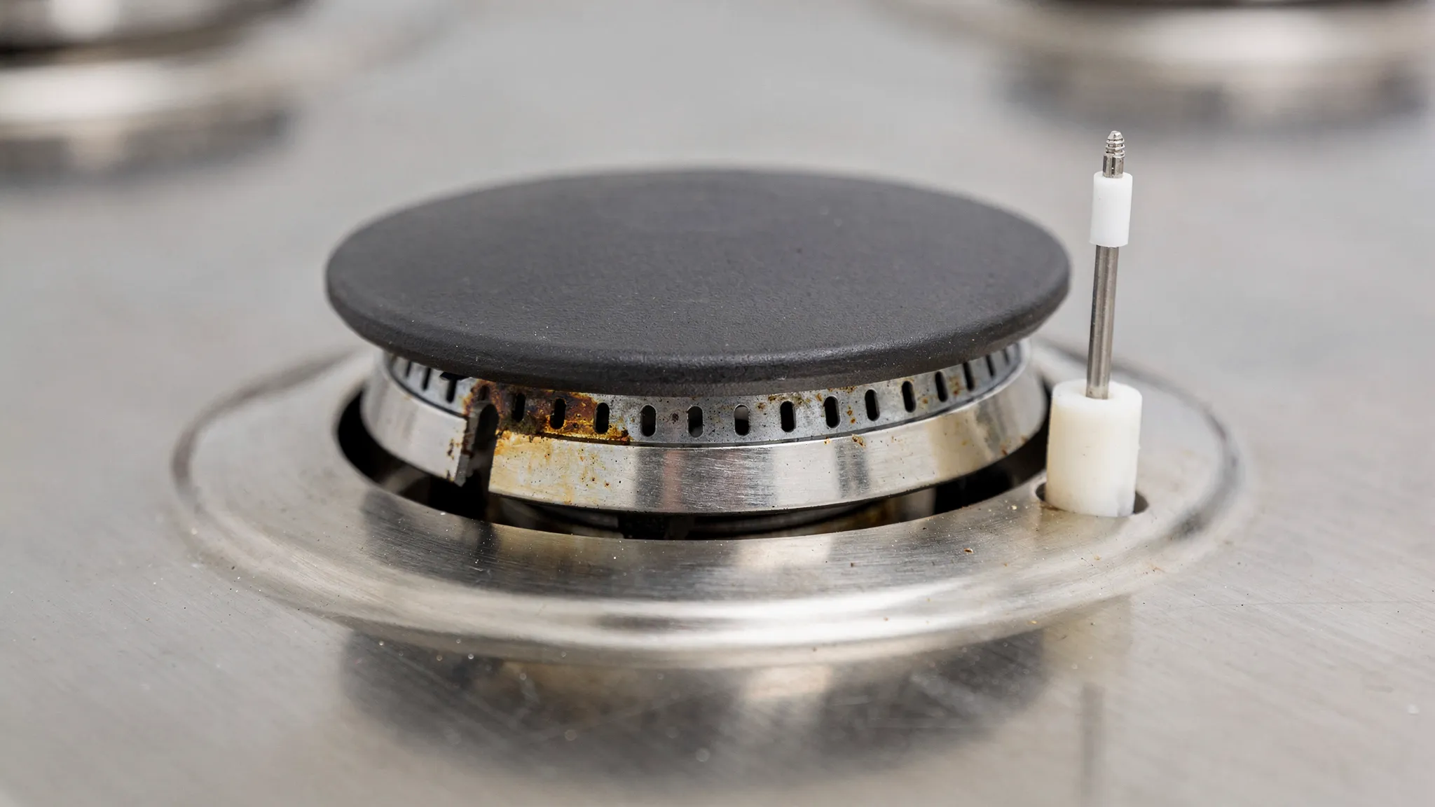 Close-up of a sealed gas stovetop burner with the grate removed, showing the burner cap and burner head, small gas ports around the ring, and a white ceramic spark electrode positioned near the edge.