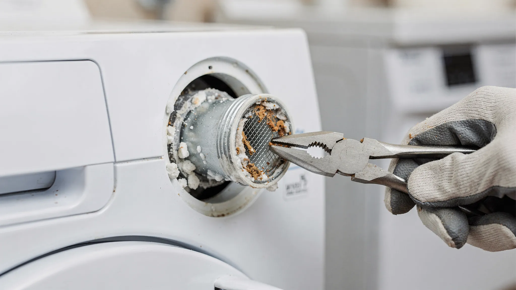 Close-up view of a washing machine water inlet connection showing a mesh filter screen partially clogged with mineral buildup and sediment, with a hand holding needle-nose pliers nearby to remove the screen safely.