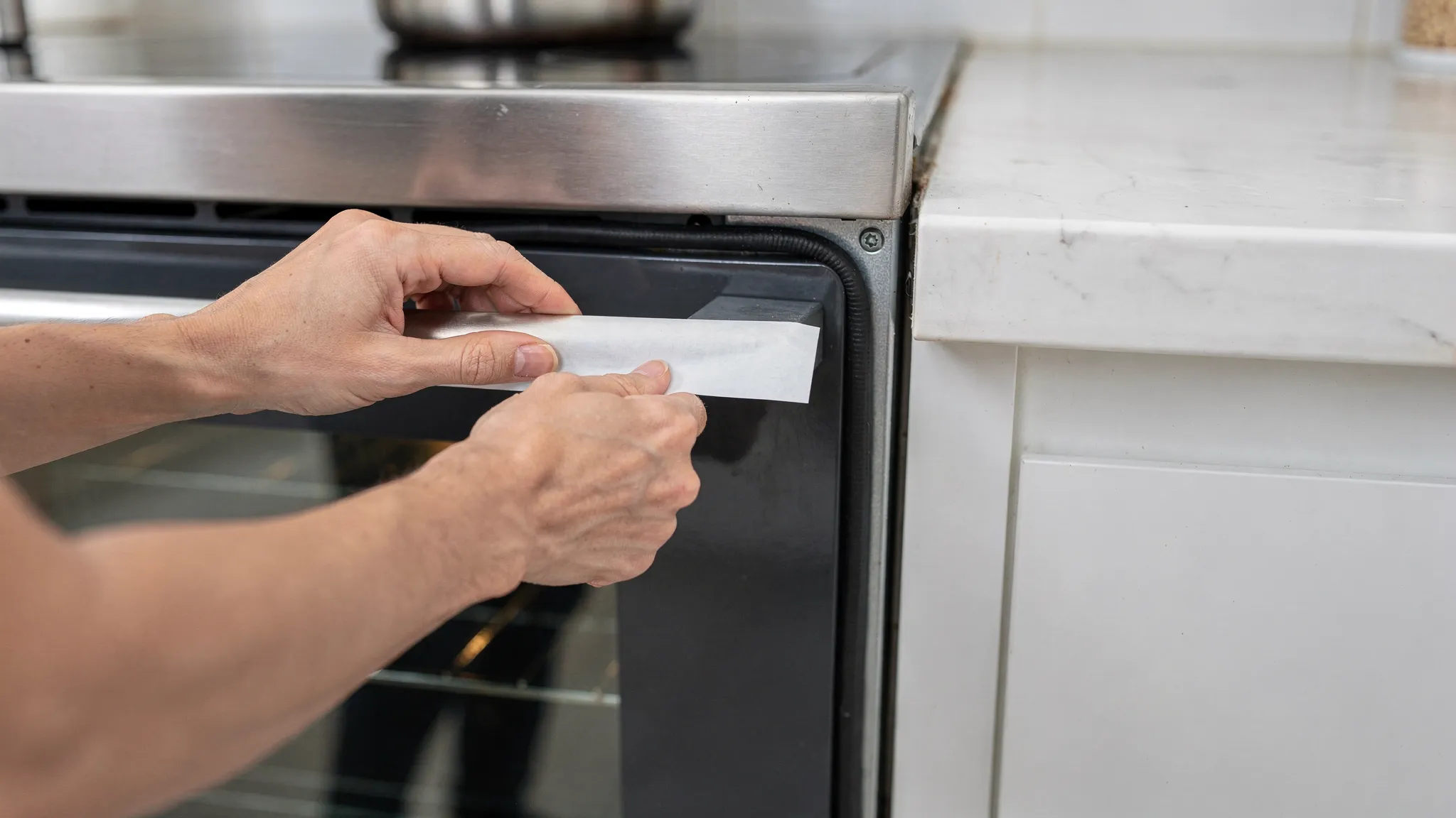 Hands performing a simple “paper test” on an oven door, with a strip of paper held between the door and the oven frame to check gasket sealing resistance.
