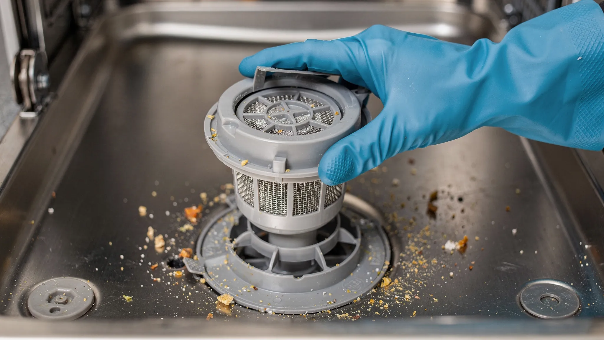 A close-up of a dishwasher tub bottom with the lower rack removed, showing a twist-lock filter being lifted out, light food debris around the sump area, and a gloved hand holding the filter for cleaning.