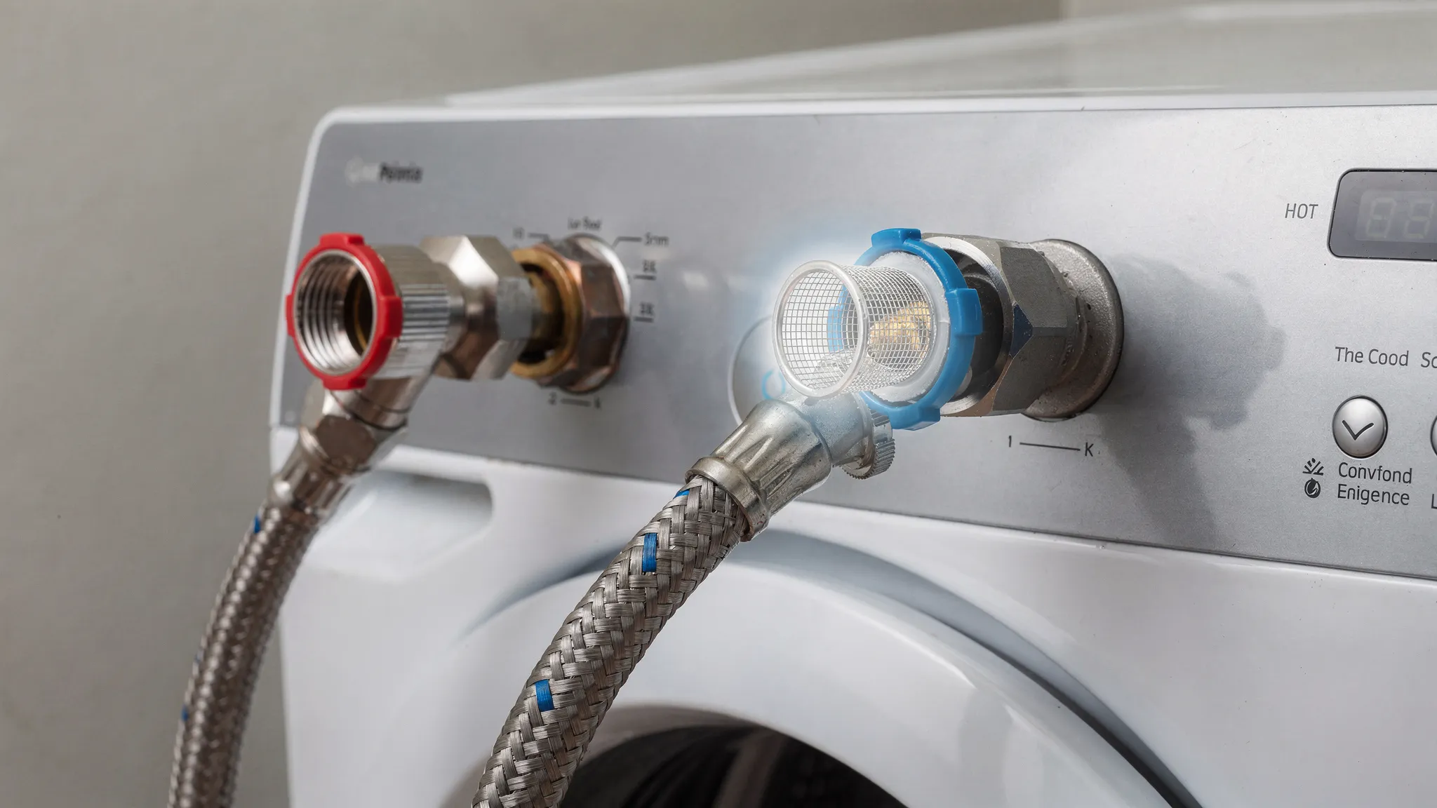 A close-up of the back of a washing machine showing hot and cold water supply valves, braided inlet hoses, and the threaded hose connections at the washer, with a small mesh inlet screen highlighted near the inlet port.