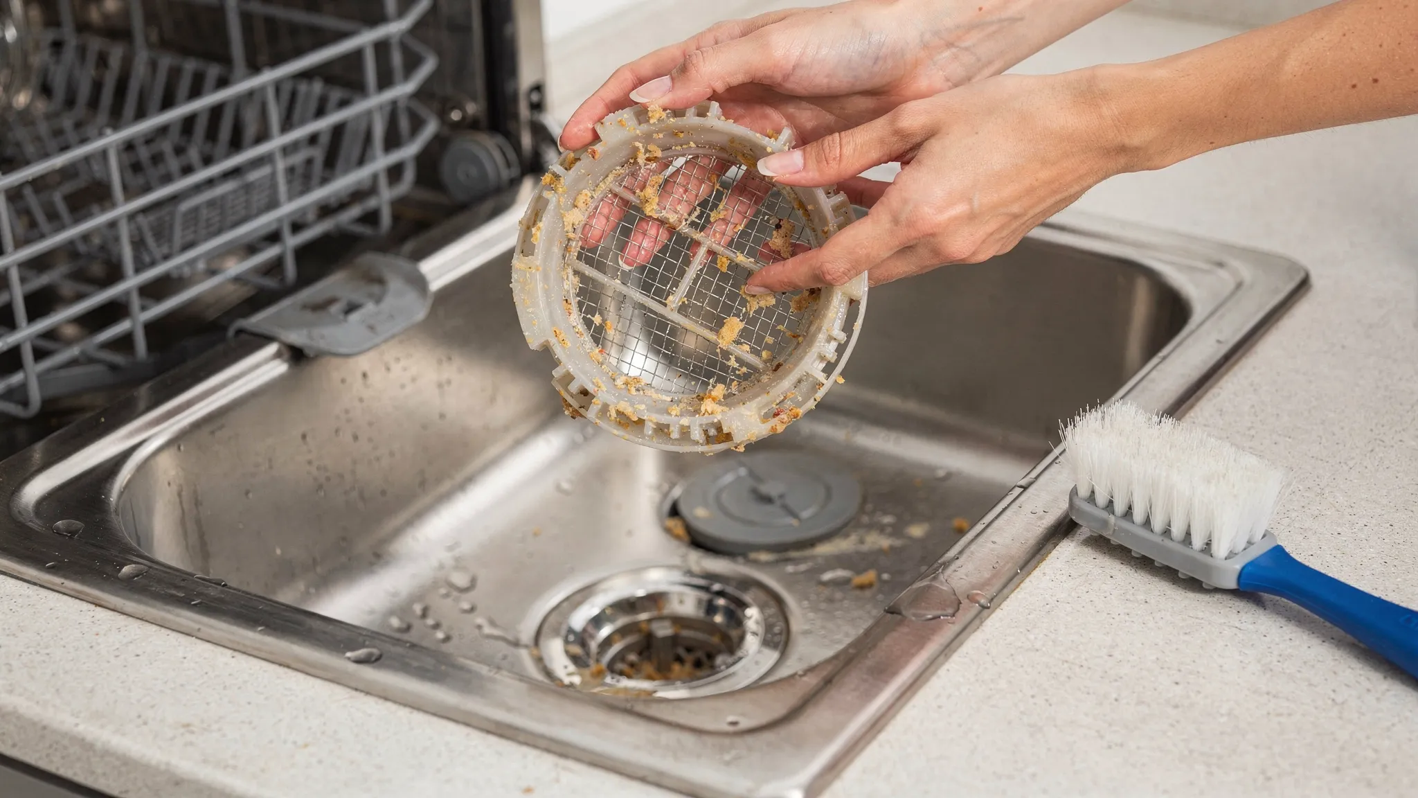 A homeowner’s hands removing a dishwasher bottom rack filter assembly over a sink, with visible food debris in the filter screen and a soft brush nearby for cleaning.
