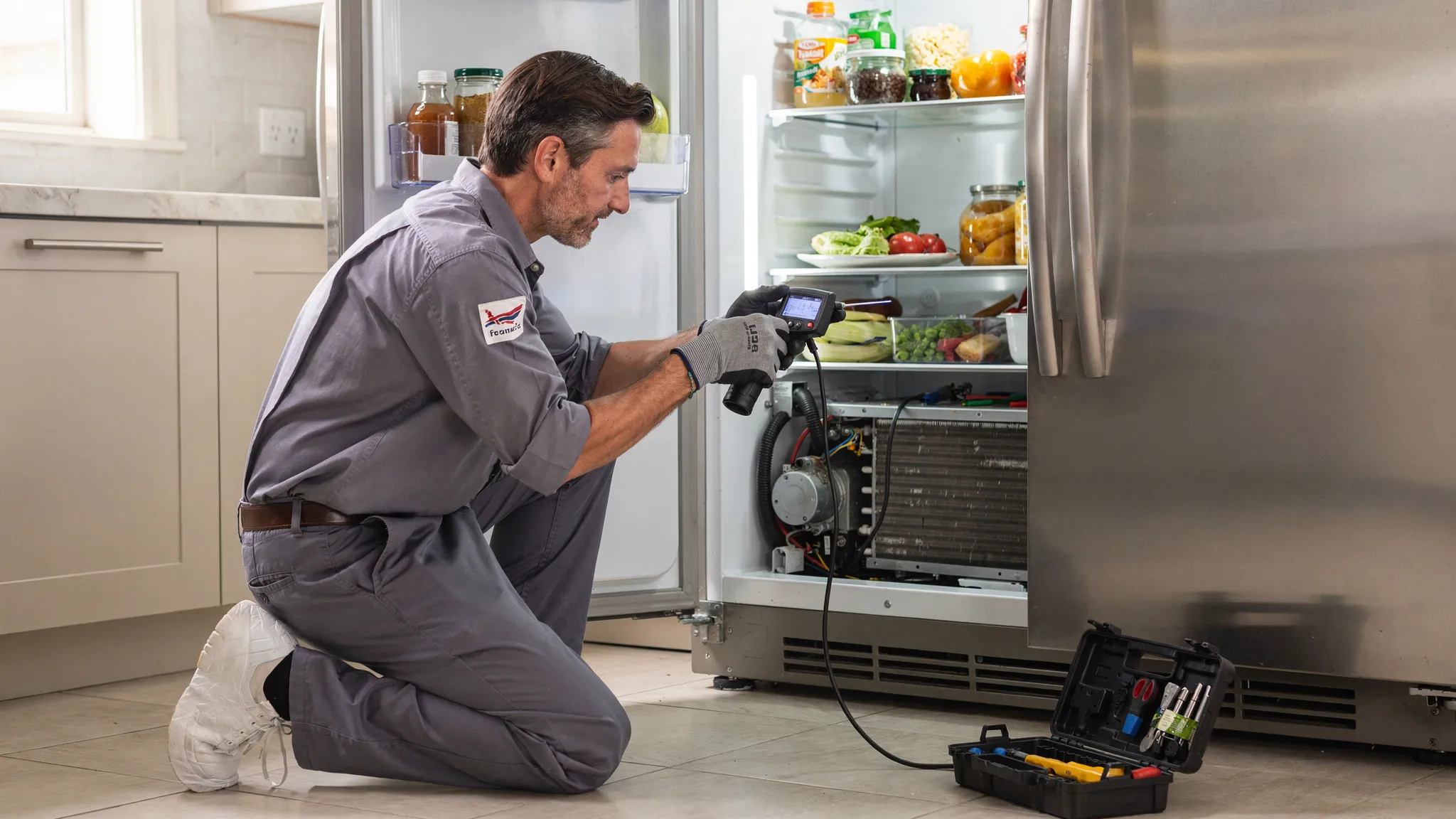 A refrigerator technician kneeling beside an open refrigerator, using a handheld thermometer and inspecting the condenser coil area with basic tools; the kitchen is tidy and well-lit, and the technician wears gloves and protective shoe covers.