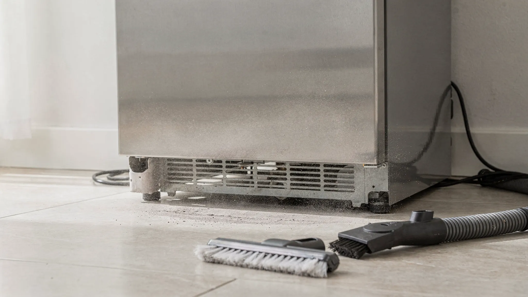 A simple scene showing a refrigerator pulled slightly away from the wall with visible clearance space behind it, a soft brush and vacuum attachment on the floor, and light dust being cleaned from a vented lower grille area.