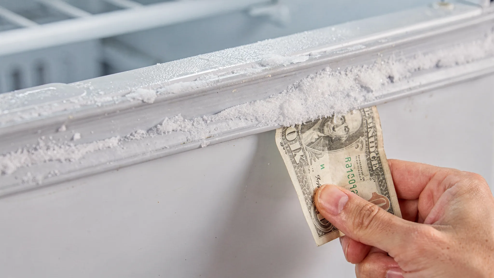 Close-up of a freezer door gasket with visible frost along the edge, plus a hand performing a “dollar-bill test” at the corner of the seal to check for leaks.