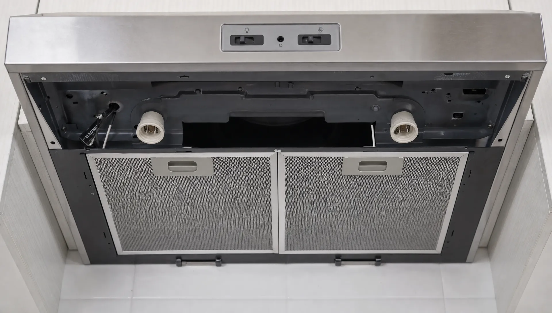 A close view of a range hood with the light cover removed, showing the bulb sockets, control switch area, and grease filters during a safe power-off inspection.